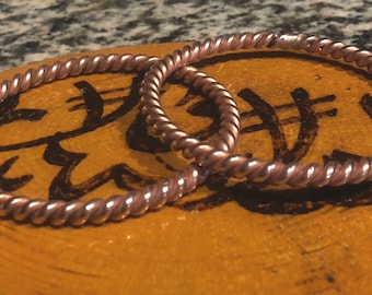 May include: Two copper wire bracelets with a braided design. The bracelets are on a wooden surface with black markings.