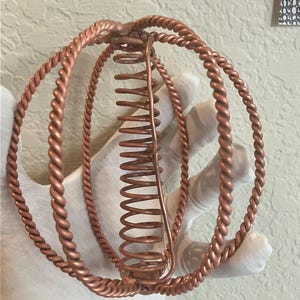 Peut inclure: Une sculpture artisanale de couleur cuivrée, composée de trois anneaux en fil torsadé et d'un élément central en forme de ressort. Les anneaux sont disposés en forme sphérique, le ressort s'étendant au centre. La sculpture est tenue par une main gantée.