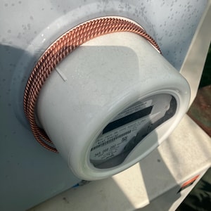 May include: Close-up of a white electrical meter with a copper-colored coil wrapped around it. The meter has a digital display and several labels with text. The coil is made of tightly wound wire.