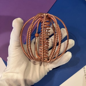 May include: A spherical sculpture constructed from multiple interlocking copper-colored twisted wire rings. The object features a central vertical coil and a small wooden bead attached to a smooth copper wire frame, creating a complex geometric design.