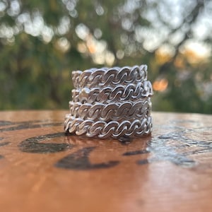 May include: A stack of four silver-colored rings with a chain-link design. The rings are stacked on a wooden surface, with a blurred green and brown background.