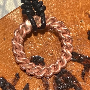 May include: A copper-colored, braided ring pendant with a black leather cord. The pendant is made of intertwined metal links, forming a circular shape. The background is a wooden surface with dark markings.