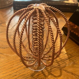 Könnte beinhalten: Kupferfarbenes Deko-Objekt mit spiralförmiger Mitte und schlingenartigen, seilartigen Armen. Das Objekt steht auf einem klaren, kreisförmigen Sockel. Das Design erinnert an ein stilisiertes Seeungeheuer oder eine Pflanzenform.
