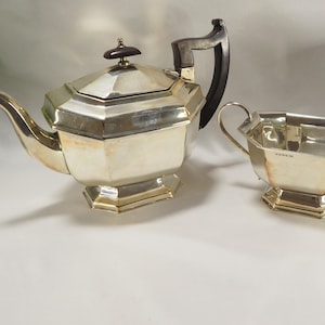 Könnte beinhalten: Eine silberne Teekanne mit einem dunkelbraunen Holzgriff, eine silberne Zuckerdose und drei weiße Porzellantassen. Die Teekanne und die Zuckerdose haben ein geometrisches Design.