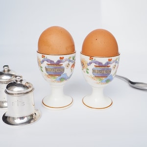 May include: Two brown eggs in white ceramic egg cups with blue and gold floral designs. The cups are on a white surface with two silver salt and pepper shakers and a silver spoon.