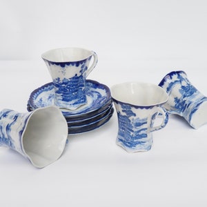 Op de afbeelding: Een set van zes witte porseleinen kopjes en schotels met een blauw-wit landschapsontwerp. De kopjes hebben een unieke, golvende vorm en de schotels hebben een geschulpte rand. De kopjes zijn ongeveer 7,6 cm hoog.