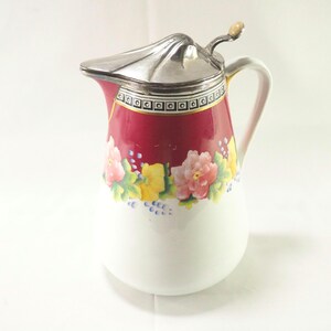 May include: A white ceramic pitcher with a red band and floral design. The pitcher has a silver lid with a small handle.