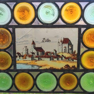 May include: Stained glass window with a central rectangular panel depicting a town scene with a church and castle, surrounded by circular glass pieces in shades of yellow, green, and orange. The word "Volk" is visible in the image.