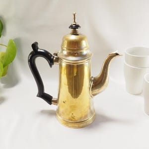 May include: A gold-tone coffee pot with a black handle and a black knob on the lid. The pot is sitting on a white surface.