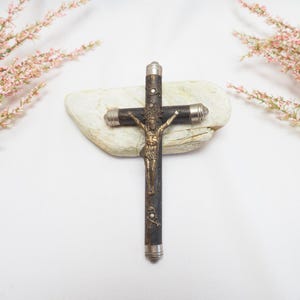 May include: A detailed crucifix featuring a bronze Jesus figure on a dark wooden cross with silver detailing. The cross rests on a light-coloured stone, with pink floral sprigs in the background.