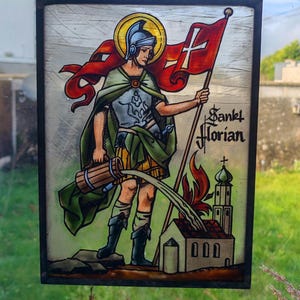 May include: Stained glass panel depicting Saint Florian, holding a flag with a white cross and a water container. The saint is dressed in armor and a green cape, with a halo. The text "Sankt Florian" is visible.