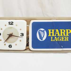 Puede incluir: Una combinación vintage de reloj y letrero publicitario. El reloj tiene una esfera cuadrada blanca con números y manecillas negras. El letrero presenta un fondo azul con las palabras "HARP LAGER" en amarillo y un logotipo de arpa.