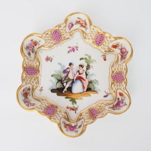Könnte beinhalten: Eine weiße Porzellanschale mit gewelltem Rand und goldener Verzierung. Die Schale zeigt eine bemalte Szene eines Paares in einer Gartenlandschaft, umgeben von Blumen und Blättern. Die Schale ist mit rosa und goldenen Akzenten verziert.