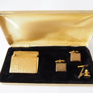 Può includere: Un set regalo dorato in una scatola foderata in velluto. Il set include un accendino dorato, un paio di gemelli e un fermaglio per cravatta.