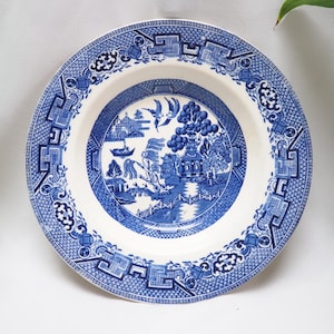 Op de afbeelding: Een ronde, wit en blauwe keramische plaat met een klassiek Willow-patroon. De plaat heeft een centrale landschapsweergave met een brug, bomen en vogels, omgeven door een decoratieve rand. De plaat heeft een diameter van ongeveer 25 cm.