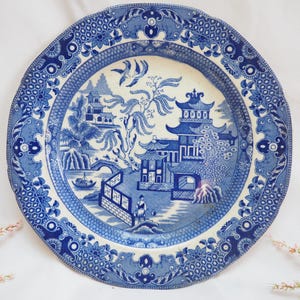May include: A blue and white ceramic plate with a detailed Willow pattern design. The plate features a scalloped edge and a central scene of a pagoda, bridge, and figures, surrounded by floral and geometric borders.