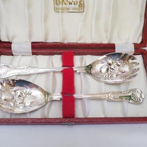 Könnte beinhalten: Ein Set aus zwei silbernen Servierlöffeln in einer roten Samtbox. Die Löffel haben ein verziertes Design mit Frucht- und Blumenmotiven. Die Box trägt den Text "Brown's Sheraton  L'Horset" auf dem Deckel.