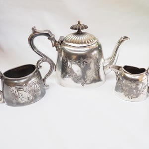 Puede incluir: Un juego de té plateado, que incluye una tetera, una jarra de crema y un azucarero, se exhibe sobre una superficie blanca. La tetera tiene una tapa y un pico decorativos. La jarra de crema y el azucarero tienen asas. Cada pieza tiene un diseño en relieve.