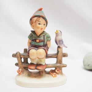 May include: A ceramic figurine of a seated boy in traditional Bavarian attire, perched on a wooden fence. A small bird sits beside him. The figurine has a cream-colored base and is decorated with muted colors.