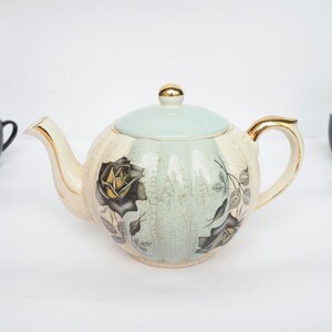 Op de afbeelding: Een witte theepot met een lichtblauw deksel en gouden accenten. De theepot heeft een bloemmotief met zwarte rozen en gouden bladeren.