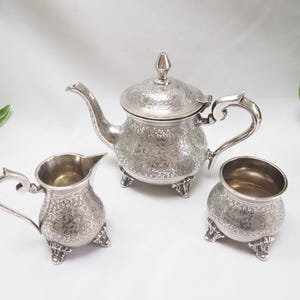 May include: A silver tea set with an ornate design. The set includes a teapot, creamer, and sugar bowl.