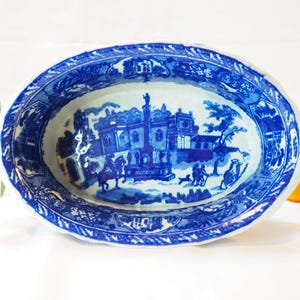 May include: Oval blue and white porcelain serving dish with a detailed landscape scene. The dish has a scalloped edge and a central design depicting buildings, figures, and trees. The colour palette is primarily blue and white.