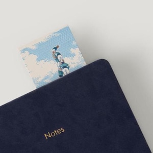 Puede incluir: Un cuaderno azul marino oscuro con la palabra Notes grabada en oro en la portada. Un marcapáginas está insertado en el cuaderno, mostrando una ilustración de cielo azul claro con nubes blancas y un grupo de figuras formando una torre humana.