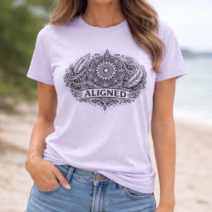 Puede incluir: Camiseta de color lila claro con un diseño gráfico negro. El diseño incluye plumas, un mandala circular y la palabra "ALIGNED". La camiseta está hecha de un material suave.