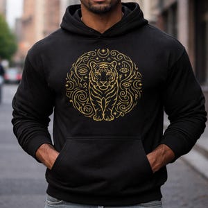 May include: Black hoodie featuring a gold tiger design within a circular pattern of stars, eyes, and swirls. The hoodie has a front pocket and a hood.