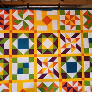 May include: A vibrant quilt with a geometric design. The quilt displays squares with diverse patterns in orange, green, purple, yellow, and white. The quilt is suspended against a blue backdrop.