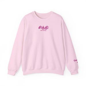 Peut inclure: Un sweat-shirt rose clair à manches longues. Le devant présente un texte magenta, avec les mots "சித்தி" au-dessus de "est 2025". La manche porte le mot "The" en magenta. Le sweat-shirt est en matière douce.