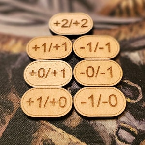 Könnte beinhalten: Sieben Holzspielsteine mit graviertem Text. Die ovalen Spielsteine zeigen numerische Werte wie +2/+2, +1/+1, -1/-1, +0/+1, -0/-1, +1/+0 und -1/-0. Die Spielsteine sind hellbraun.