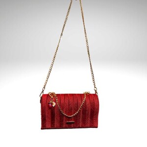 May include: A red handbag with a gold chain strap. The bag features a textured, vertical striped pattern and a small gold charm with a flower and gem. The chain is attached to the bag with gold hardware.