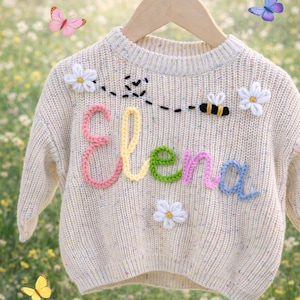 Puede incluir: Jersey de punto color crema con el nombre "Elena" en hilo colorido. El jersey presenta margaritas bordadas, una abeja y mariposas. El jersey está colgado de una percha de madera sobre un fondo floral borroso.