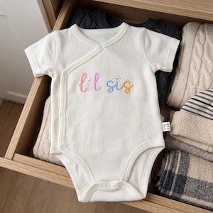 May include: A cream-colored, short-sleeved, ribbed baby bodysuit featuring a wrap-style front with snap closures. The chest displays the embroidered text "lil sis" in colorful cursive lettering, accented with small white daisy flower details.