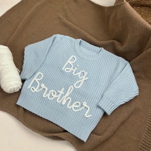 May include: A light blue knit sweater with the words "Big Brother" embroidered in white. The sweater is displayed on a brown blanket, with a ball of white yarn and a woven basket in the background. The sweater is designed for a baby or toddler.