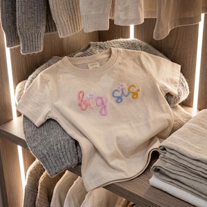 以下が含まれることがあります： 筆記体で「big sis」と刺繍されたベージュの半袖Tシャツ。「big」の文字はピンクのグラデーションで、「sis」は青と黄色で縫い付けられています。小さな刺繍の花が文字のアクセントになっています。