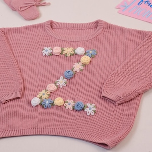Könnte beinhalten: Ein altrosa Strickpullover mit einem floralen "Z"-Design. Der Pullover hat einen Rundhalsausschnitt und lange Ärmel. Ein Paar passende Hausschuhe und ein rosa Wimpel mit Text sind ebenfalls sichtbar. Kinderkleidung.