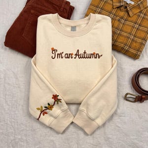 Può includere: Una felpa girocollo color crema con il testo ricamato "I'm an Autumn" in marrone, accompagnato da piccoli dettagli floreali arancioni e gialli sulla manica sinistra. Il capo è mostrato insieme a pantaloni in velluto a coste marrone e una camicia a quadri.