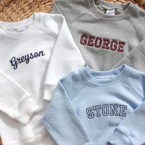 Può includere: Tre felpe girocollo in bianco, grigio e azzurro, ciascuna ricamata con un nome. La felpa bianca dice "Greyson" in corsivo. La felpa grigia dice "GEORGE" in lettere maiuscole bordeaux. La felpa azzurra dice "STONE" in lettere maiuscole blu navy.