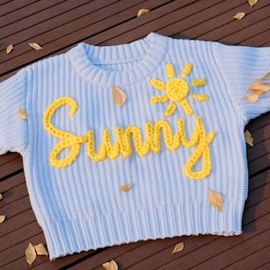 May include: Light blue knit sweater with the word "Sunny" in yellow script and a yellow sun graphic. The sweater is short-sleeved and has a ribbed hem and cuffs. The background is a wooden surface with scattered leaves.