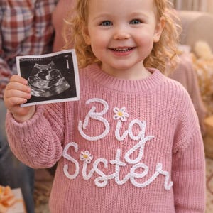 May include: A young girl smiles, holding an ultrasound image. She wears a pink sweater with "Big Sister" embroidered in white, accented with small flower details. The image suggests a pregnancy announcement.