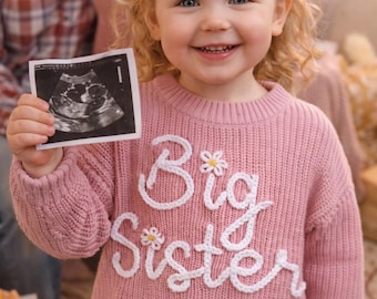 Könnte beinhalten: Ein lächelndes junges Mädchen hält ein Ultraschallbild. Sie trägt einen rosa Pullover mit weißer Stickerei "Big Sister", verziert mit kleinen Blumen. Das Bild deutet auf eine Schwangerschaftsankündigung hin.
