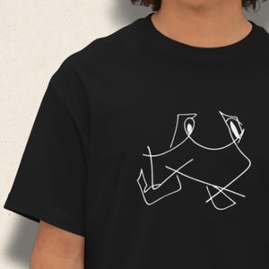 Op de afbeelding: Zwart t-shirt met een witte abstracte lijntekening op de voorkant. De afbeelding lijkt een gestileerd gezicht of figuur te zijn. Het shirt heeft korte mouwen en een ronde hals.