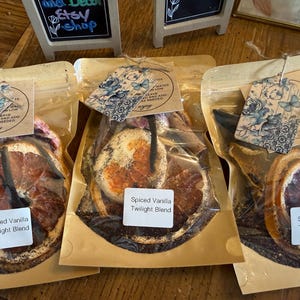 May include: Three clear plastic bags filled with dried fruit slices and spices, each with a tag featuring a floral design. The bags have labels that read "Spiced Vanilla Twilight Blend". The background includes a wooden surface and small chalkboard signs.