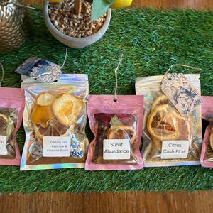 May include: Five small pink pouches containing dried citrus slices and herbs, each labeled with a different intention: Wealth and Wisdom, Fortune Fire, Sunlit Abundance, Citrus Cash Flow, and Money Magnet. The pouches are on a green surface.