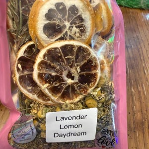 May include: A clear plastic bag filled with dried lemon slices, herbs, and chamomile flowers. The bag has a pink border and a label that reads "Lavender Lemon Daydream". The bag is sealed and ready for use.