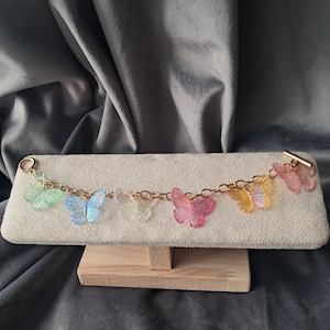 May include: A gold chain bracelet with seven translucent butterfly charms in various colours. The butterflies are green, blue, clear, pink, and yellow. The bracelet is displayed on a cream-coloured jewellery display stand.