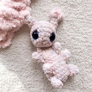 May include: A light pink crocheted plush toy featuring a soft, fuzzy texture, two prominent antennae with tufted ends, and two large, round, glossy black eyes. The creature has a rounded head and a segmented body with four small limbs.