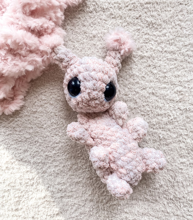 May include: A light pink crocheted plush toy featuring a soft, fuzzy texture, two prominent antennae with tufted ends, and two large, round, glossy black eyes. The creature has a rounded head and a segmented body with four small limbs.